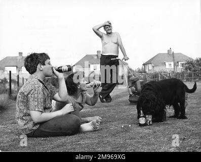 Un dimanche de banlieue dans le jardin à l'arrière. Mère et fils se gardent au frais avec des bouteilles de boisson, tandis que le chien s'entête dans un verre de limonade des Caraïbes. Papa, surchauffé, se laisse une pause de la tonte de la pelouse. Banque D'Images