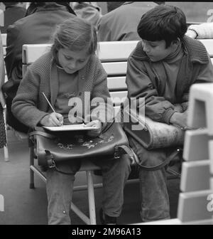 Une fille et un garçon avec leurs cartables d'école, assis sur un banc. La fille écrit quelque chose dans son carnet, tandis que le garçon regarde. Banque D'Images