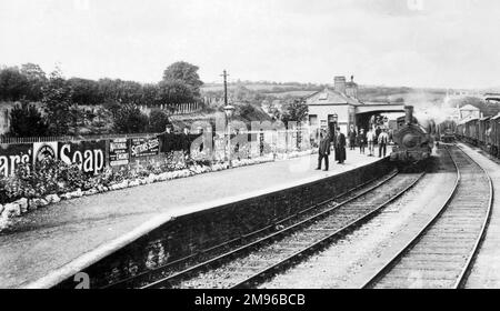 Vue d'une plate-forme de la gare de Cardigan sur le Great Western Railway, Cardiganshire, au sud du pays de Galles, avec des parterres de fleurs et des annonces en plaques pour Pears SOAP et Sutton Seeds. Un train à vapeur attend à la plate-forme, et la plupart des gens, passagers et personnel, regardent directement la caméra. Banque D'Images