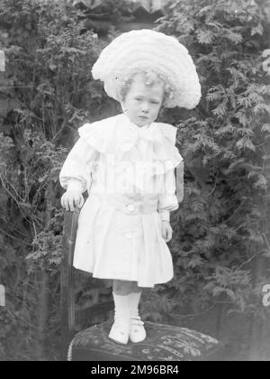 Une petite fille édouardienne debout sur une chaise dans un jardin, au Moyen-Galles, portant un énorme chapeau blanc, une robe blanche, des chaussettes et des chaussures blanches. Ses cheveux sont en ringlets blonds. Banque D'Images