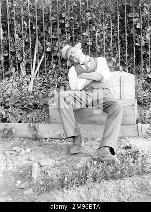 Un homme dans un chapeau de paille et des manches courtes, endormi dans un jardin, au centre du pays de Galles. Il semble avoir fait du jardinage, car il a laissé ses tondeuses devant les rampes derrière lui. Le jardinage est si épuisant! Banque D'Images