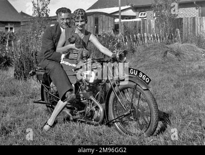 Un couple assis sur une moto dans un jardin. La femme tient un spaniel sur ses genoux. Banque D'Images