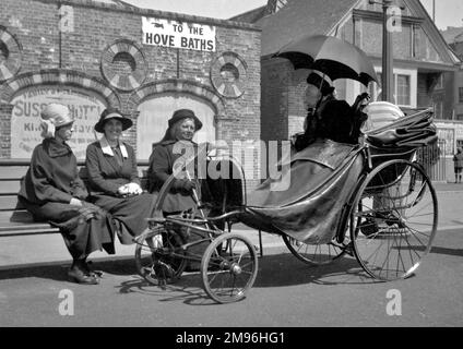 Scène de rue près des bains de Hove à Sussex. Trois femmes s'assoient sur un banc, près d'une femme âgée dans une chaise de bain à quatre roues élaborée, ombragée du soleil par un parapluie noir. Banque D'Images