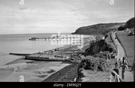 Une station balnéaire non identifiée avec un quai et une jetée, et des personnes marchant le long d'un chemin de falaise sur la droite. Banque D'Images