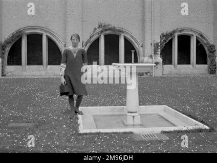 Une jeune femme debout près d'une fontaine au milieu d'une pelouse pleine de pâquerettes. Banque D'Images