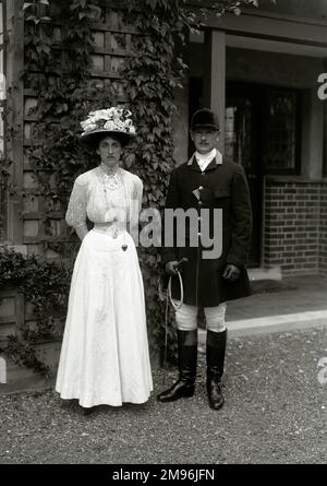 Une femme en robe typiquement édouardienne et chapeau, posant pour sa photo à l'extérieur d'une maison avec un homme en tenue de chasse. Banque D'Images