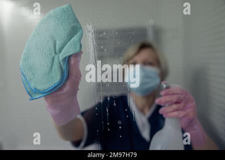 La femme de chambre principale dans le masque nettoie le miroir dans la salle de bains de l'hôtel en vaporisant du détergent sur la surface Banque D'Images