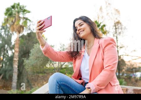 Une jeune fille de latina prend un selfie avec son téléphone cellulaire assis dans un escalier dans un parc extérieur. Banque D'Images