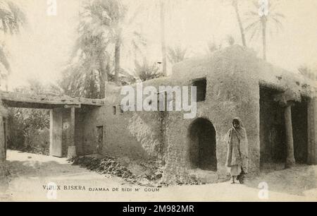 Un homme âgé se trouve à côté d'une maison faite de matériaux argileux, au coin d'une rue dans le vieux Biskra. Banque D'Images