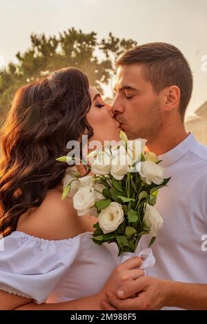 Un jeune couple s'embrasse en gros plan. Portrait des amoureux des baisers. Guy et fille en vêtements blancs et avec bouquet blanc de roses Banque D'Images