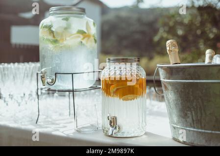 Seau à glace rempli de bouteilles de vin et de champagne à côté des verres et deux distributeurs d'eau remplis d'oranges et de citron sur une table de jardin Banque D'Images