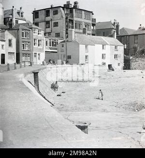 L'une des grandes séries d'études de St Ives, en Cornouailles. Au bout du quai et au début de Quay Street. Deux femmes sur la plage du port avec leur vélo et un petit garçon jouant avec son scooter. Près d'eux est un chariot de bateau presque enterré avec des roues de fer. Au bout du quai, deux personnes s'assoient sur un banc pour discuter. Un autre vélo est garé contre le pignon des cottages. Banque D'Images