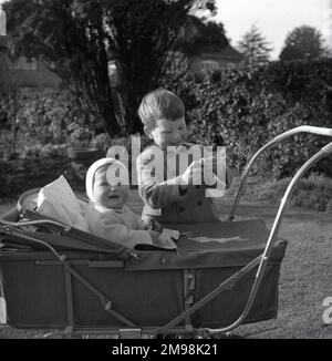 1960s, historique, à l'extérieur d'un jardin, un jeune garçon debout à côté de son frère qui, avec un coussin derrière lui, est assis dans un pram à cadre métallique de l'époque, Angleterre, Royaume-Uni. Ce type de chariot porte-voitures pour bébé ou bébé était de la même conception que le pram traditionnel à quatre roues construit par autocar, où l'enfant est face à la personne poussant le pram, mais était une version plus légère et plus petite, ayant un cadre qui signifie qu'il se repliait quand il n'est pas utilisé. Banque D'Images