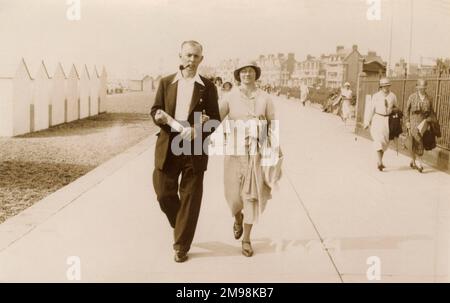 Couple de vacances promenade le long de la promenade à Felixstowe, Suffolk. Les badges sur sa veste indiquent peut-être qu'il est un cheminwayman et que sa femme porte quelques romans à un moment donné les heures de résidence dans son transat... Banque D'Images