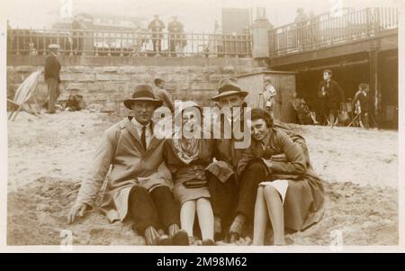 Scène typique de l'été britannique - deux couples se sont enveloppés de chaleur sur la plage en le bravant dans des manteaux et des chapeaux. Peut-être pris à Blackpool. Banque D'Images