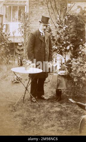 Homme élégant (peut-être un peu trop habillé ?!) et sa jeune fille dans le jardin, qui porte un manteau et des bottes très élégants. Ils sont debout (curieusement) dans un jardin plutôt effroné qui donne sur le dos! Banque D'Images