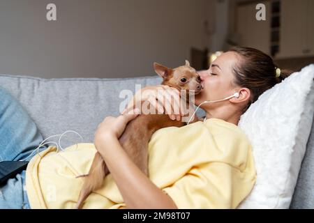 Brunette femme se reposant à la maison avec son petit chien allongé sur un canapé embrassant son chien et écoutant de la musique à l'aide d'un casque et d'un téléphone portable. Banque D'Images