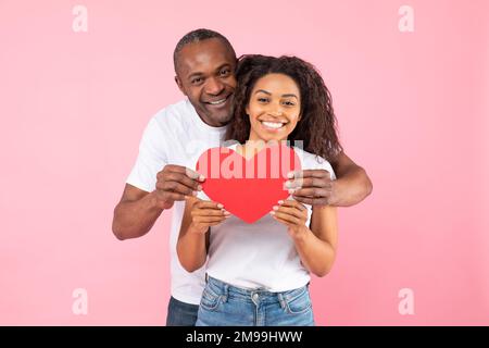 Amoureux heureux noir épouses tenant coeur en papier, homme embrassant la femme brom dos et souriant à l'appareil photo, fond rose Banque D'Images