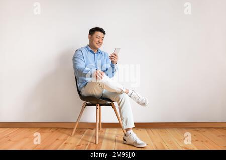 Homme asiatique d'âge moyen enthousiaste utilisant un smartphone, la mise en réseau sur les réseaux sociaux, assis sur une chaise au-dessus du mur blanc du studio Banque D'Images