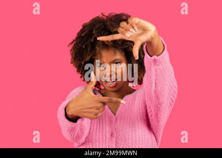 Bonne jeune femme afro-américaine curly avec des bretelles montrer le signe, faire le cadre avec les mains, regarder à travers elle Banque D'Images