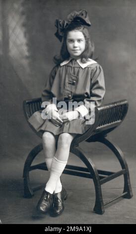 Jolie jeune fille avec un énorme noeud en velours dans ses cheveux assis pour un portrait photographique studio sur un élégant tabouret incurvé siège. Banque D'Images