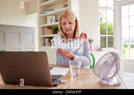 Femme mature ménopausique souffrant de douleurs articulaires au coude travaillant sur ordinateur portable à la maison Banque D'Images