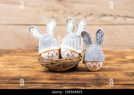 Trois oeufs de Pâques aux visages drôles et chapeaux crochetés gris aux oreilles de lapin dans un nid décoratif de sisal sur fond de bois. Concept joyeuses Pâques. Banque D'Images