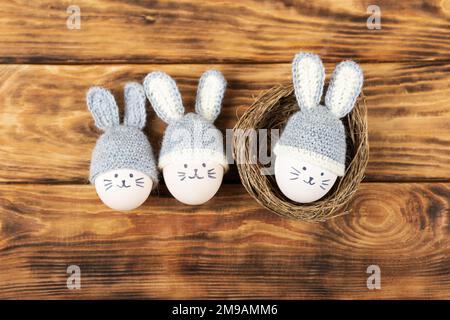 Trois oeufs de Pâques aux visages drôles et chapeaux crochetés gris aux oreilles de lapin dans un nid décoratif de sisal sur fond de bois. Concept joyeuses Pâques. Banque D'Images