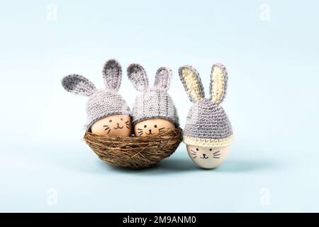 Trois oeufs de Pâques aux visages drôles et chapeaux crochetés gris aux oreilles de lapin dans un nid décoratif de sisal sur fond bleu. Concept joyeuses Pâques. G Banque D'Images