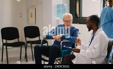Médecin généraliste en consultation avec la personne âgée en fauteuil roulant, remplissant les rapports médicaux dans le hall de l'hôpital. Patient souffrant d'une incapacité chronique dans la salle d'attente discutant avec un médecin. Banque D'Images