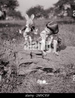 1920S PETITE FILLE PENCHÉE SUR DES ROCHERS AVEC PELUCHE LAPIN DE PÂQUES PETER LAPIN À CÔTÉ D'ELLE REGARDANT VERS LE BAS SUR LE NID DES OEUFS DE PÂQUES - E13 HAR001 HARS HAR001 À L'ANCIENNE Banque D'Images