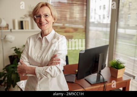 Portrait d'une femme à la maturité, pigiste dans des lunettes debout sur le fond du bureau à domicile Banque D'Images