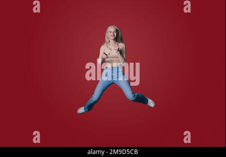 Portrait de la jeune femme souriante posant jumpant écartant les jambes regardant la caméra isolée sur couleur rouge mur arrière-plan studio Banque D'Images