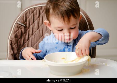 Un enfant drôle mange une pomme râpée avec sa bouche pleine tout en étant assis sur une chaise de cuisine. Bébé garçon affamé, il a de la nourriture dans sa bouche, de l'humour. Enfant âgé Banque D'Images