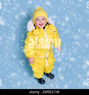 Bébé heureux en hiver vêtements de neige sur fond bleu studio. Un enfant dans une combinaison jaune chaud avec capuche. Enfant âgé d'un an et de cinq mois Banque D'Images