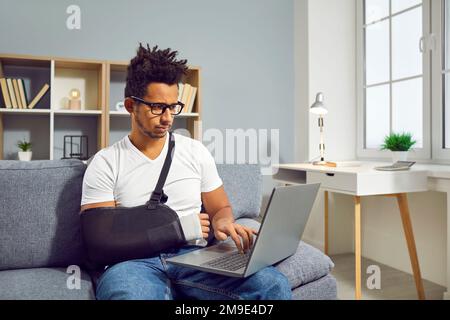 Homme avec un poignet cassé dans une élingue de bras assis sur un canapé à la maison et travaillant sur un ordinateur portable Banque D'Images