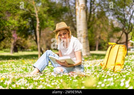 Une jeune fille blonde dans un chapeau lisant un livre au printemps dans un parc de la ville, des vacances à côté de la nature et à côté de pâquerettes Banque D'Images