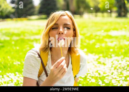 Une femme blonde souriante soufflant sur la plante de pissenlit, portant un chapeau et des lunettes de soleil assis sur l'herbe au printemps à côté de pâquerettes dans un parc dans le Banque D'Images