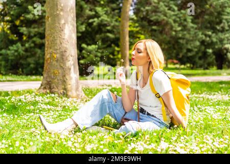 Une femme blonde soufflant sur l'usine de pissenlits dans un parc de la ville, portant un chapeau et des lunettes de soleil assis sur l'herbe au printemps à côté des pâquerettes Banque D'Images