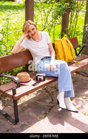 Portrait d'une blonde touriste en vacances d'été assis sur un banc dans un parc, portant un chapeau, un appareil photo, sac à dos jaune Banque D'Images