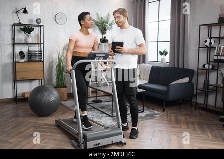 Portrait d'une femme afro-américaine portant des vêtements de sport s'entraîner avec un tapis roulant et parler à un entraîneur d'homme caucasien. Entraînement de fitness à domicile deux personnes sur la piste de course à pied stationnaire intelligente. Banque D'Images