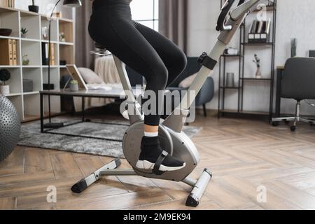 Coupe courte des jambes des athlètes féminins portant des vêtements de sport et utilisant un vélo d'exercice. Home fitness entraînement femme sportive entraînement sur vélo d'appartement élégant à l'intérieur. Banque D'Images