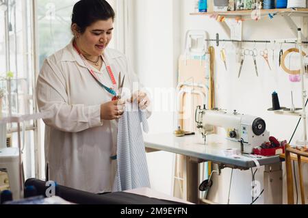 Une jeune femme caucasienne se tenant dans son atelier calme et pensant comment couper un morceau de tissu pour une conception de vêtements avec des ciseaux, debout à côté de la Banque D'Images