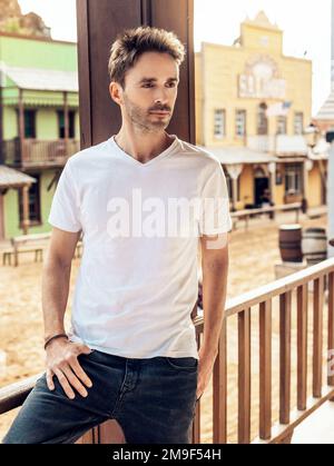 Un jeune homme beau à barbe porte un t-shirt blanc vide et se tient sur le fond de la rue ouest de la ville. Modèle vertical. Banque D'Images