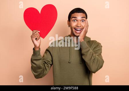 Photo de jeune romantique drôle gars excités positive toucher joue rouge papier amour symbole comme son partenaire isolé sur fond beige couleur Banque D'Images