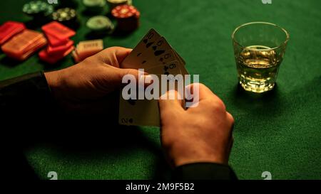 Main de gros plan tenant des cartes de poker, avec un verre de whisky et des jetons de poker dispersés sur la table verte texturée. Banque D'Images