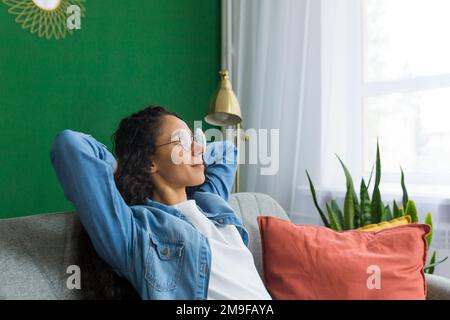 Belle femme hispanique à la maison de près avec les yeux fermés se détendant sur le canapé, femme avec les mains derrière la tête souriant dans le salon. Banque D'Images