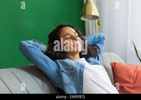 Belle femme hispanique à la maison de près avec les yeux fermés se détendant sur le canapé, femme avec les mains derrière la tête souriant dans le salon. Banque D'Images