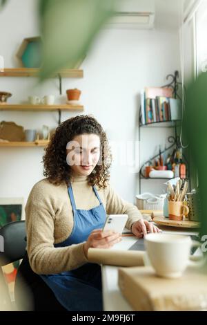 Femme céramiste lisant un message texte sur son smartphone. Femme d'affaires créative gérant un magasin avec une collection de produits céramiques. Entrée jeune Banque D'Images