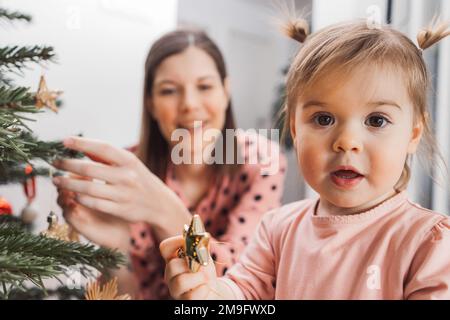 Portrait de bébé fille curieux avec deux petites queues de cheval tenant un ornement, maman floue dans l'arrière-plan décorant l'arbre de Noël Banque D'Images